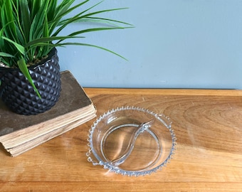 May include: A clear glass divided serving dish with a beaded edge and a handle. The dish is circular with a central divider, perfect for serving snacks or condiments. It sits on a wooden surface, with a potted plant in the background.