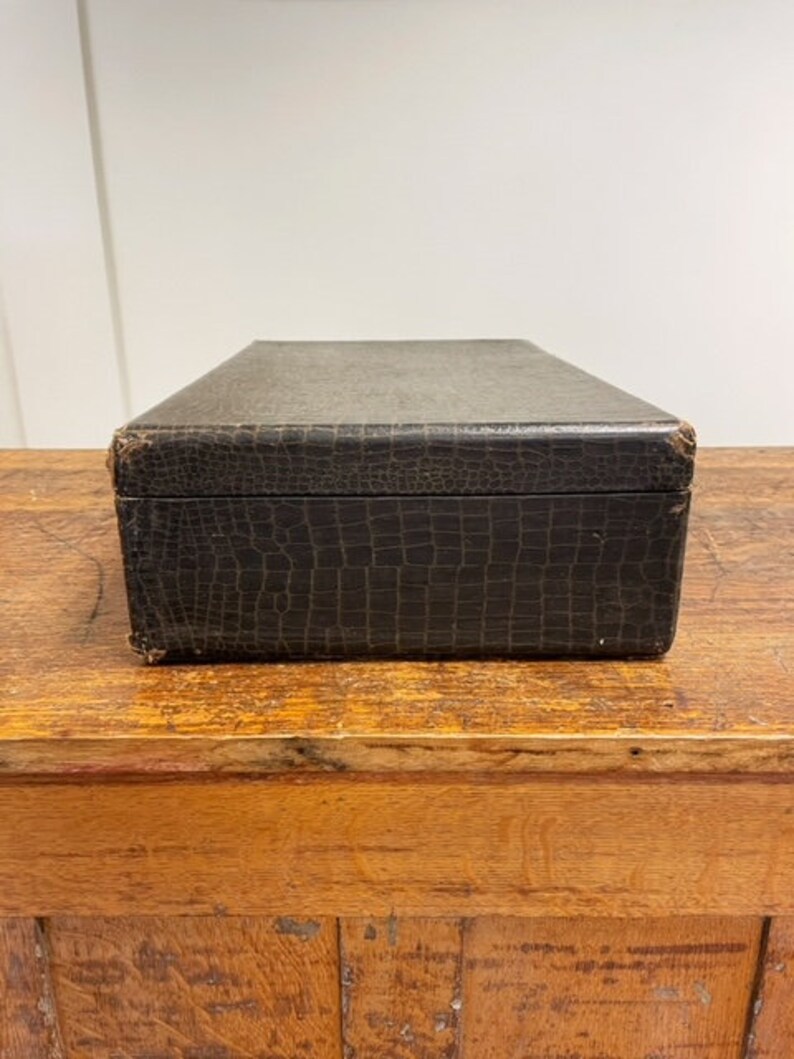 May include: A dark brown leather box with a crocodile-embossed pattern. The box is sitting on a wooden surface.