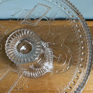 May include: Clear glass serving platter with embossed floral and geometric patterns. The platter features a central, raised, fluted handle and decorative edging. The design includes a horseshoe and rectangular shapes. The glass is transparent, allowing the surface beneath to be visible.