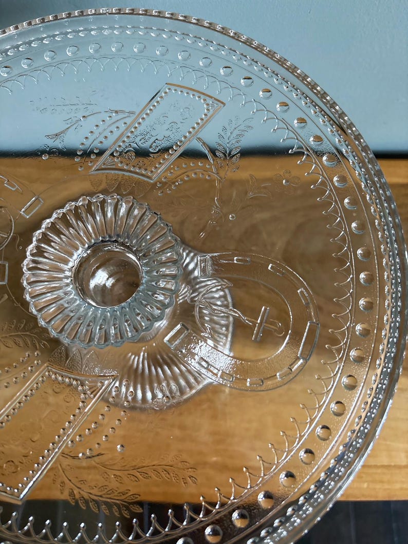 May include: Clear glass serving platter with embossed floral and geometric patterns. The platter features a central, raised, fluted handle and decorative edging. The design includes a horseshoe and rectangular shapes. The glass is transparent, allowing the surface beneath to be visible.