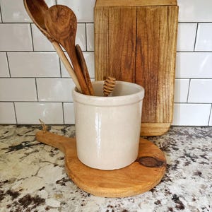 May include: A cream-colored ceramic crock holds several wooden spoons and a honey dipper. The crock sits on a round wooden cutting board, while a rectangular wooden cutting board stands upright behind it, showcasing natural wood grain patterns.
