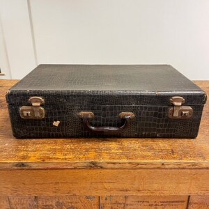 May include: A vintage black suitcase with a textured leather-like finish and brass hardware. The suitcase has a brown leather handle and is closed.