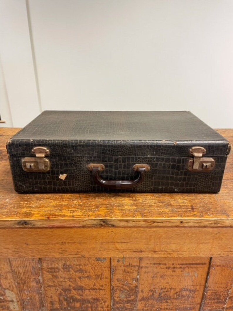 May include: A vintage black suitcase with a textured leather-like finish and brass hardware. The suitcase has a brown leather handle and is closed.