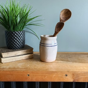 May include: A cylindrical beige stoneware crock featuring two horizontal blue stripes near the rim. Two wooden spoons with visible grain patterns are placed inside the crock. The crock has a smooth finish and a slightly flared top edge.