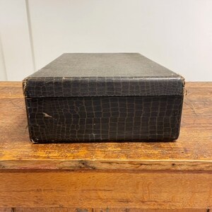 May include: A vintage, black, rectangular box with a crocodile-embossed leather exterior. The box is sitting on a wooden surface.