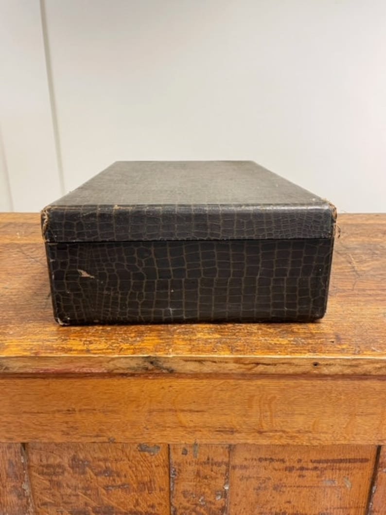 May include: A vintage, black, rectangular box with a crocodile-embossed leather exterior. The box is sitting on a wooden surface.