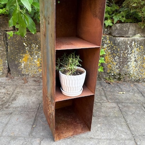 May include: A tall, rectangular, rusted metal shelf unit with three shelves. A small white pot with a plant sits on the middle shelf. The unit has a weathered, aged appearance, with shades of brown and gray.