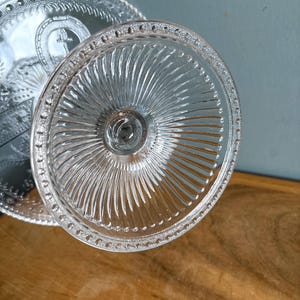 May include: Clear glass cake stand with a ridged base and a detailed, beaded edge. The stand's surface features a cross design. The glass reflects light, creating a sparkling effect. A classic piece for serving desserts.