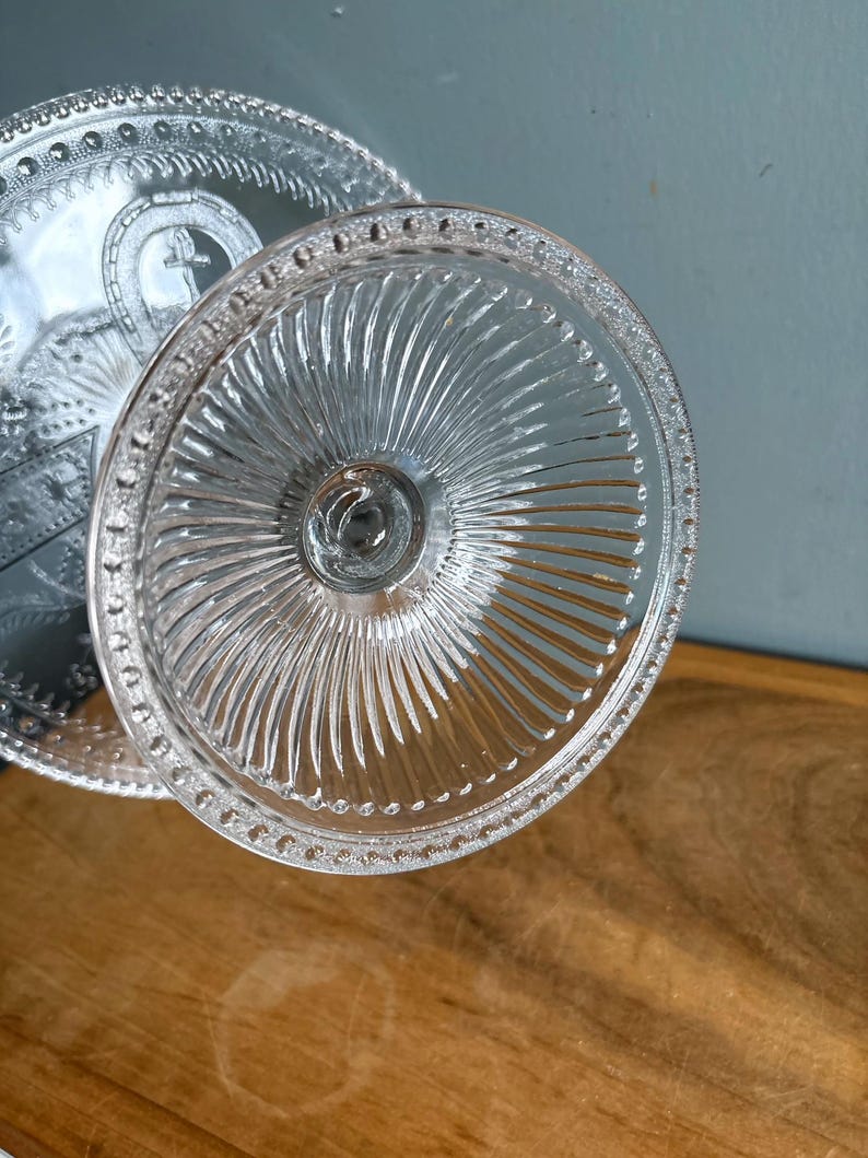 May include: Clear glass cake stand with a ridged base and a detailed, beaded edge. The stand's surface features a cross design. The glass reflects light, creating a sparkling effect. A classic piece for serving desserts.