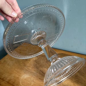 May include: Clear glass cake stand with a round top and a decorative etched design. The stand has a pedestal base and a detailed rim. The cake stand is on a wooden surface.