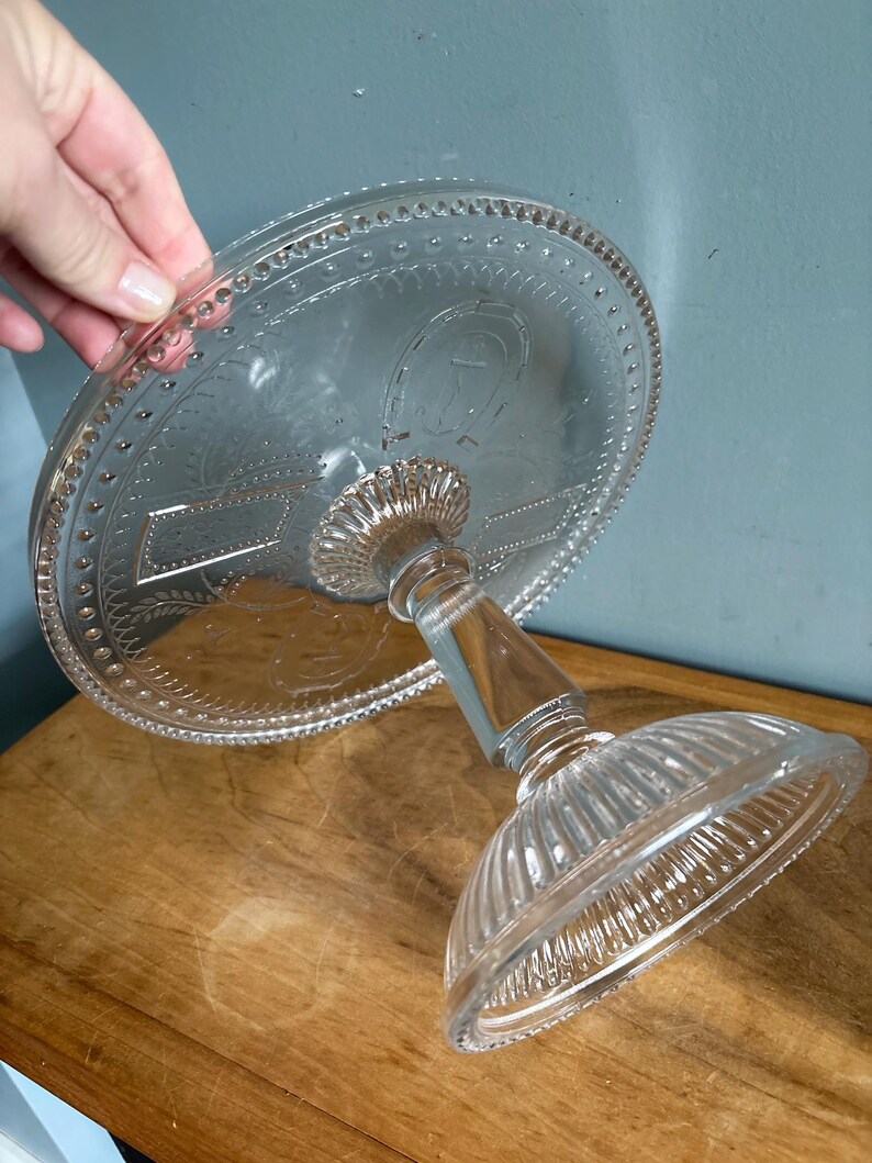 May include: Clear glass cake stand with a round top and a decorative etched design. The stand has a pedestal base and a detailed rim. The cake stand is on a wooden surface.