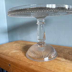 May include: A clear glass cake stand with a round top and a pedestal base. The top edge has a beaded design. The base has vertical ridges. The stand is on a wooden surface.