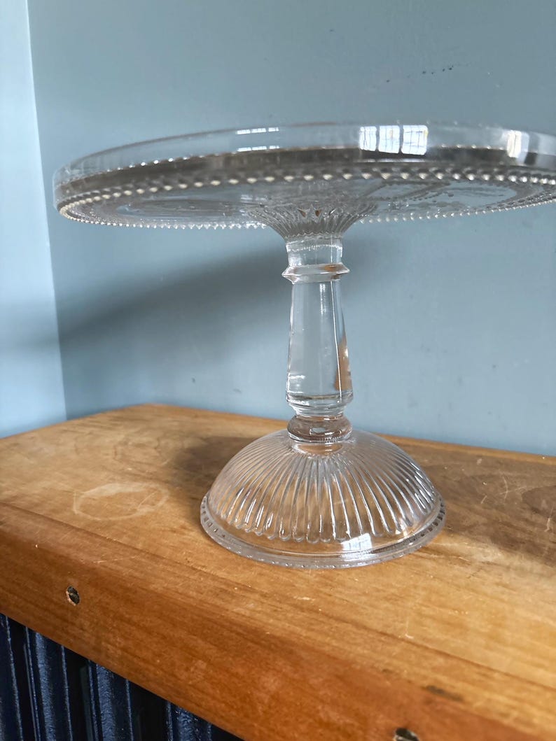 May include: A clear glass cake stand with a round top and a pedestal base. The top edge has a beaded design. The base has vertical ridges. The stand is on a wooden surface.