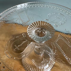 May include: Clear glass serving tray with a central pedestal and etched designs. The tray has a circular base with a raised centre for holding items. Decorative elements include horseshoe and rectangular patterns.
