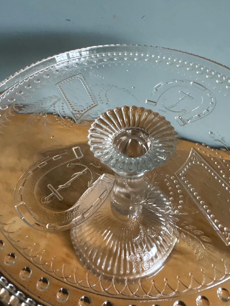 May include: Clear glass serving tray with a central pedestal and etched designs. The tray has a circular base with a raised centre for holding items. Decorative elements include horseshoe and rectangular patterns.