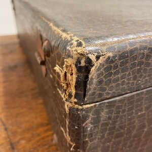 May include: A vintage, brown leather-bound box with a worn, textured surface. The box has a damaged corner, revealing the fabric lining underneath.