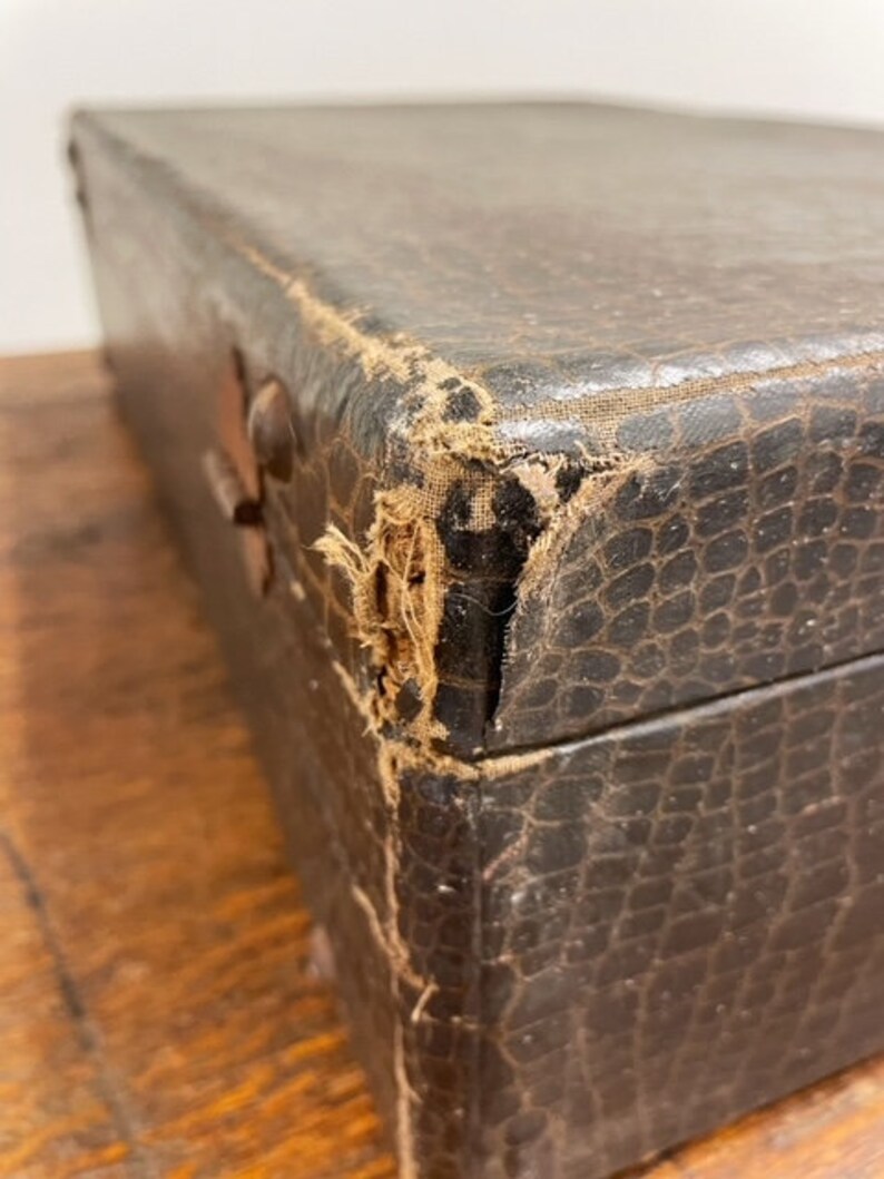 May include: A vintage, brown leather-bound box with a worn, textured surface. The box has a damaged corner, revealing the fabric lining underneath.