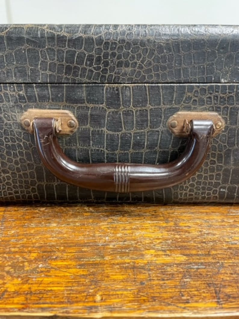 May include: A black vintage suitcase with a crocodile-embossed leather exterior and a brown plastic handle.