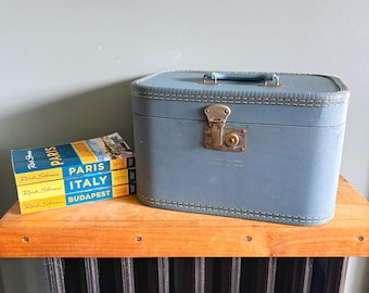 May include: A light blue rectangular hard-shell travel case with a top handle and a metal latch closure, positioned next to a stack of three travel guidebooks titled Rick Steves Paris 2015, Rick Steves Italy 2010, and Rick Steves Budapest.