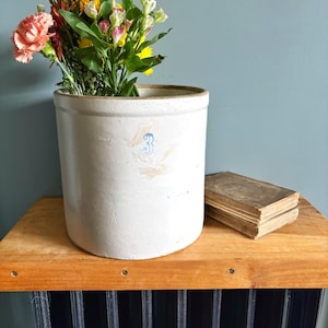 May include: A cylindrical light gray stoneware crock featuring a blue number 3 stamped on the front. The crock is filled with a colorful bouquet of fresh flowers and sits next to a weathered, vintage hardcover book with worn edges.