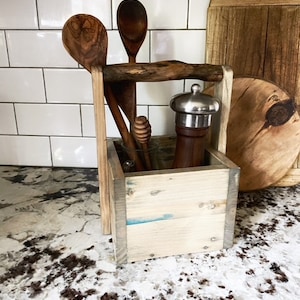 May include: A wooden utensil holder with a handle, holding wooden spoons, a pepper grinder, and a honey dipper. The holder is made of light-colored wood with a blue accent. A wooden cutting board is in the background.