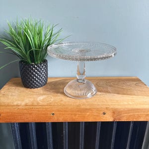 May include: A clear glass cake stand with a round top and a pedestal base. The top has a decorative pattern around the edge. The base has vertical ridges. A green plant in a black pot sits to the left.
