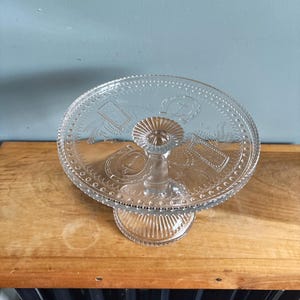 May include: A clear glass cake stand with a round, decorative top and a pedestal base. The top features embossed designs, including a horseshoe and other shapes. The stand is on a wooden surface.