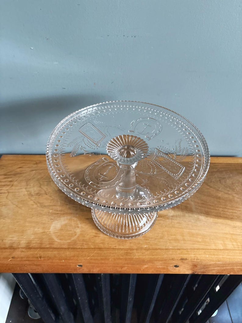 May include: A clear glass cake stand with a round, decorative top and a pedestal base. The top features embossed designs, including a horseshoe and other shapes. The stand is on a wooden surface.