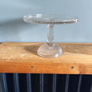 May include: A clear glass cake stand with a round top and a decorative beaded edge. The stand has a short, ribbed stem and a circular base. The stand is displayed on a wooden surface.