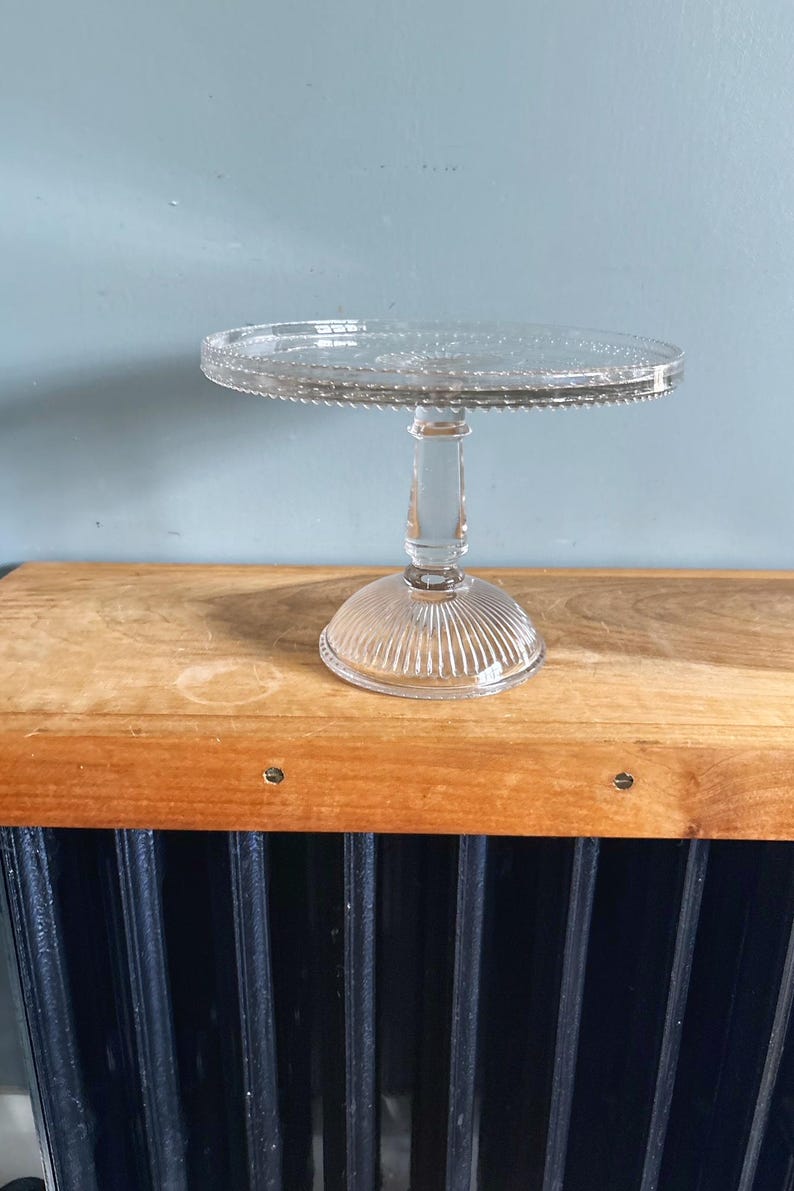 May include: A clear glass cake stand with a round top and a decorative beaded edge. The stand has a short, ribbed stem and a circular base. The stand is displayed on a wooden surface.