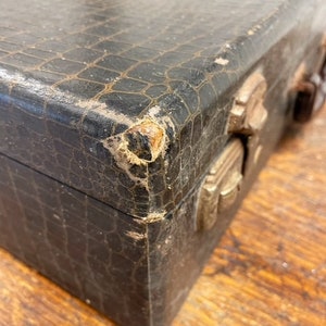 May include: A vintage, black, crocodile-embossed leather box with brass clasps and hinges. The box shows signs of wear and tear, adding to its vintage charm.