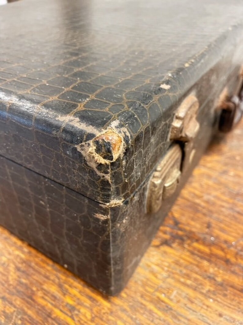 May include: A vintage, black, crocodile-embossed leather box with brass clasps and hinges. The box shows signs of wear and tear, adding to its vintage charm.