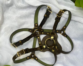 Puede incluir: Un arnés corporal de cuero verde oliva oscuro con correas ajustables, hebillas metálicas doradas, remaches y anillos circulares. El diseño incluye un panel central de cuero conectado a múltiples correas que se cruzan formando una estructura geométrica.
