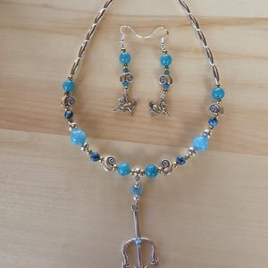 May include: A jewelry set consisting of a necklace and matching earrings. The necklace features silver-toned beads, blue round beads, small spiral shell charms, and a central silver trident pendant. The earrings feature blue beads and horse charms.