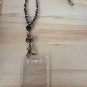May include: A beaded lanyard featuring metallic bronze-toned beads and dark speckled stone beads. The lanyard includes a bronze-colored spiral metal charm and a lobster claw clasp attached to a clear, rectangular plastic ID badge holder.