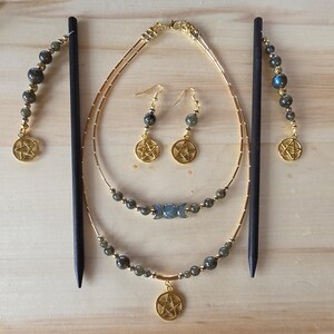 May include: A jewelry set consisting of two dark wooden hair sticks, a pair of drop earrings, and a two-strand necklace. The pieces feature gold-tone metal chains, grey labradorite beads, and gold-tone pentagram charms. The necklace includes a triple moon symbol.