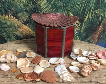 Op de afbeelding: Een rode glazen doos met een schelpdeksel. De doos is omringd door verschillende schelpen op een houten tafel.