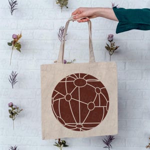 Op de afbeelding: Een beige canvas draagtas met een groot, bruin geometrisch ontwerp dat lijkt op een netwerk of globe. De tas wordt door een persoon vastgehouden, met de handvatten zichtbaar. De achtergrond is een witte bakstenen muur met bloemaccenten.