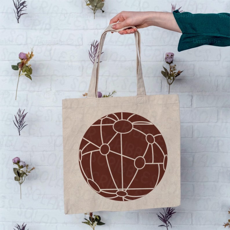 Op de afbeelding: Een beige canvas draagtas met een groot, bruin geometrisch ontwerp dat lijkt op een netwerk of globe. De tas wordt door een persoon vastgehouden, met de handvatten zichtbaar. De achtergrond is een witte bakstenen muur met bloemaccenten.