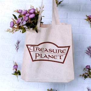 Op de afbeelding: Een beige canvas draagtas met de woorden "Treasure Planet" in een bruin, gestileerd lettertype. De tas is gevuld met paarse bloemen en groen. De tas hangt tegen een witte bakstenen muur.