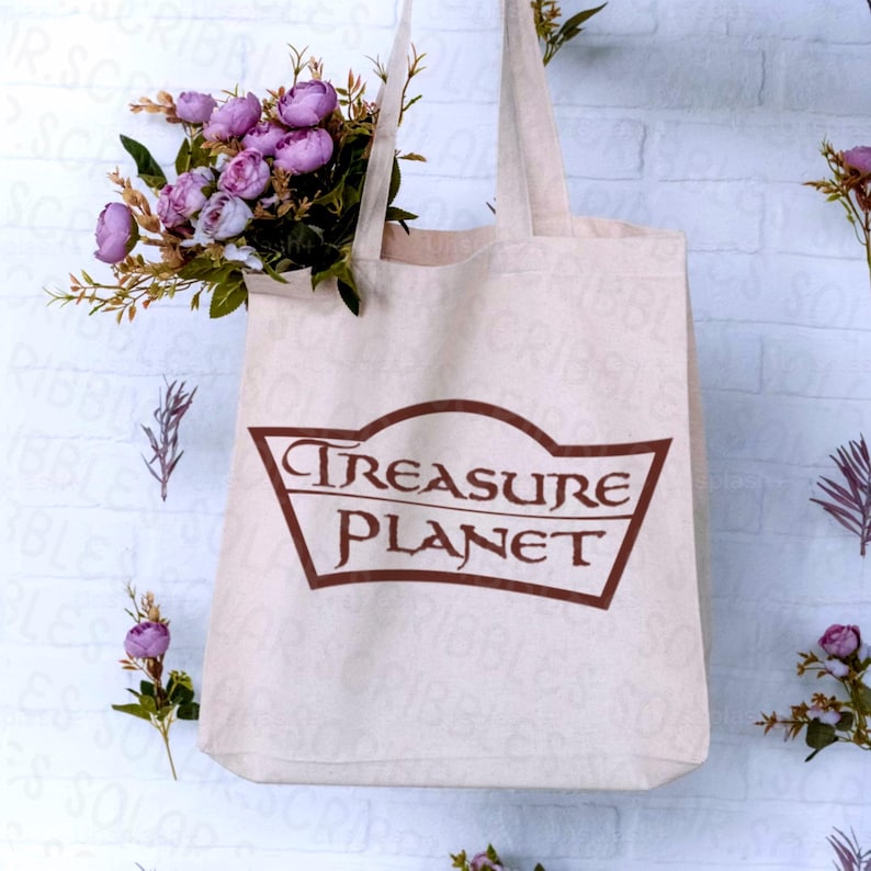 Op de afbeelding: Een beige canvas draagtas met de woorden "Treasure Planet" in een bruin, gestileerd lettertype. De tas is gevuld met paarse bloemen en groen. De tas hangt tegen een witte bakstenen muur.