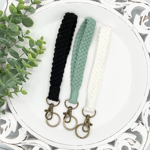 May include: Three macrame wrist keychains in black, sage green, and white, each with a bronze-colored clasp and ring. The keychains are displayed on a white, decorative plate.