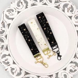 May include: Three keychains with gold star patterns on black, white, and cream fabric. The keychains have gold key rings and gold star charms.