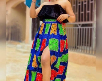 May include: A woman wearing a black strapless top and a long, colorful skirt with a high slit. The skirt has a geometric pattern in shades of blue, red, green, and yellow. She is wearing red braids and sunglasses.