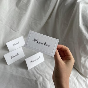 May include: White place cards with elegant black calligraphy. The cards display names such as "Hamilton," "Laurel," "Camille," and "Susannah." The cards are held against a white fabric background.