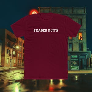 以下が含まれることがあります： 胸の中央に白い「TRADER DJO'S」の文字が入った、マルーン色のTシャツ。クルーネックのスタイルです。背景には、建物や街灯のある夜の街並みが写っています。