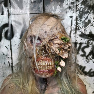 Puede incluir: Un espantoso maquillaje de zombi con dientes expuestos, carne en descomposición y un crecimiento fúngico cerca del ojo. El zombi tiene pelo largo y rubio pálido y el pecho rasgado. El fondo presenta arte de graffiti.