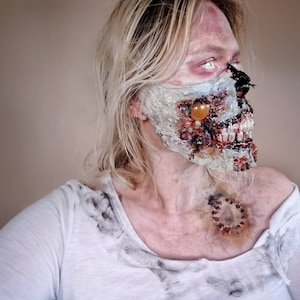 Może przedstawiać: Osoba z blond włosami i makijażem zombie. Makijaż obejmuje odsłonięte zęby, rozkładającą się twarz i ranę na szyi. Osoba ma na sobie białą koszulkę z ciemnymi plamami.