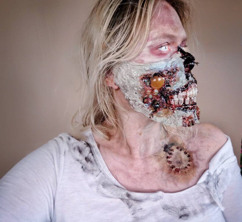 Może przedstawiać: Osoba z blond włosami i makijażem zombie. Makijaż obejmuje odsłonięte zęby, rozkładającą się twarz i ranę na szyi. Osoba ma na sobie białą koszulkę z ciemnymi plamami.
