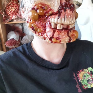 Może przedstawiać: Straszna, realistyczna maska zombie z odsłoniętymi zębami, krwią i krostami. Maska jest noszona na czarnym t-shircie z kolorową grafiką zombie pijącego herbatę i tekstem "#Brainsforte".